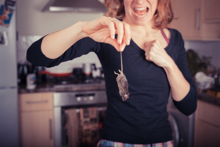 A woman in a kitchen recoils in disgust while holding a dead mouse by its tail.