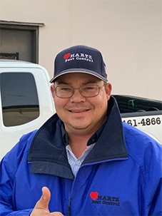 A smiling man in a Hartz Pest Control uniform gives a thumbs up in front of a company truck.