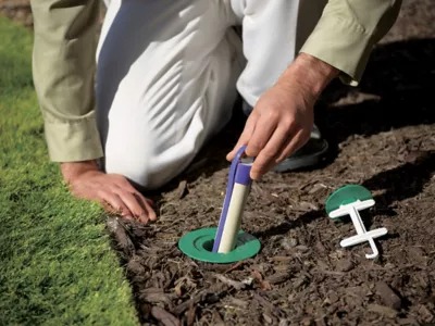 A person kneeling in a garden inserts a cylindrical device into a green base in the soil, next to a white accessory.